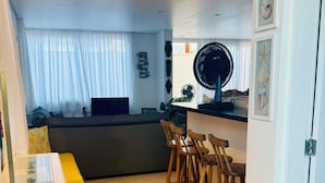 A modern living room with a sofa, dining table, and chairs. There is a large window with sheer curtains, a wall-mounted fan, and a decorative wall piece. - BEAUTIFUL HOUSE CAMBURY Gated Community 5 Bedrooms 300mts from the Beach, Camburi, S.Sebastião (SÃO SEBASTIÃO)