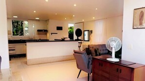 A modern kitchen with a central island, a dining area with a chair, and a wooden cabinet with a fan. - BEAUTIFUL HOUSE CAMBURY Gated Community 5 Bedrooms 300mts from the Beach, Camburi, S.Sebastião (SÃO SEBASTIÃO)
