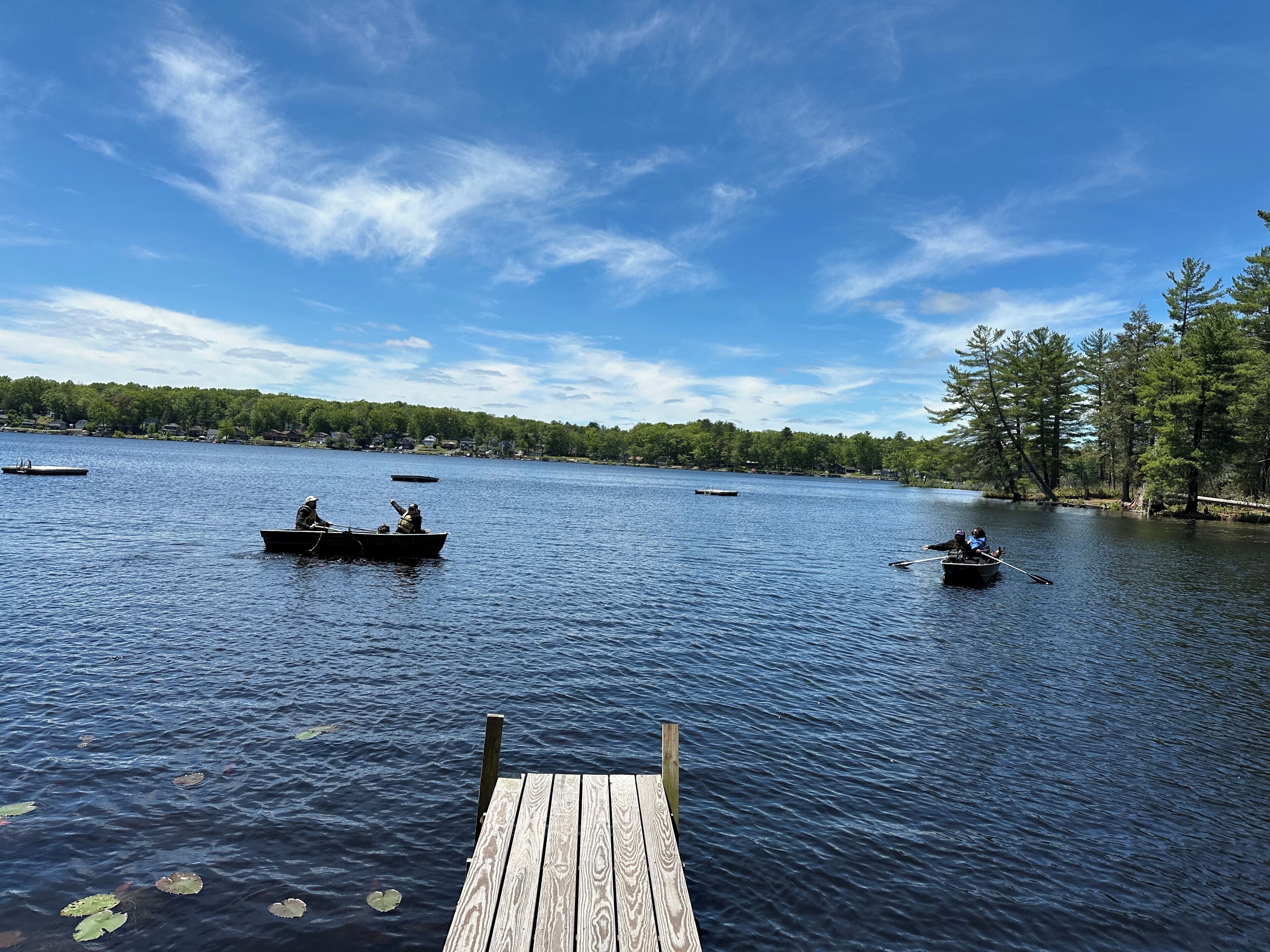 A wooden dock extending into a calm lake with several boats on the water and a forested shoreline in the background.