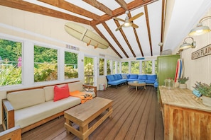 A covered outdoor seating area with wooden flooring, a ceiling fan, and a view of greenery through large windows.