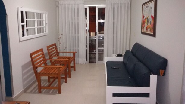 A living room with a sofa, two wooden chairs, a small table, a window with white curtains, and a door leading to another room. - Sleeps 6 people, I love this place. (Jardim Praiano)