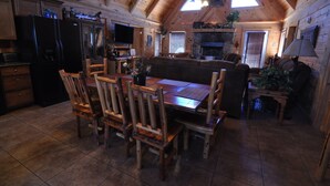 A cozy cabin interior with a wooden dining table, chairs, a sofa, a fireplace, and a kitchen area with a refrigerator and cabinets. - Wilderness Retreat Cabin- Lg Game Room, Private Hot Tub; 1 Mile to Thunder Ridge (Ridgedale)