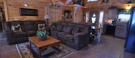 A spacious living room with a large sectional sofa, a wooden coffee table, and a fireplace. The room has wooden walls and a high ceiling with exposed beams.