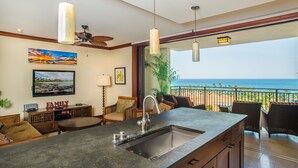 A modern kitchen with a large island, a dining area with wicker furniture, and a balcony with a view of the ocean.
