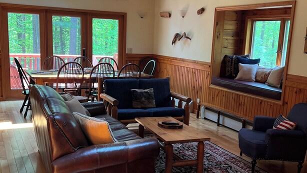 A spacious living room with a leather sofa, a coffee table, and a rug. There are multiple windows offering views of the surrounding forest. - Bright cabin retreat happily situated 7 miles from Whiteface Mt (Jay)
