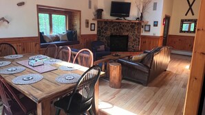 A spacious living room with a wooden dining table set for six, a stone fireplace, a television mounted above it, and a cozy seating area with a sofa and armchairs. - Bright cabin retreat happily situated 7 miles from Whiteface Mt (Jay)