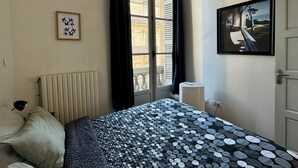 Una camera d'albergo con un letto, un termosifone, una finestra con tende, un quadro appeso al muro e una porta.