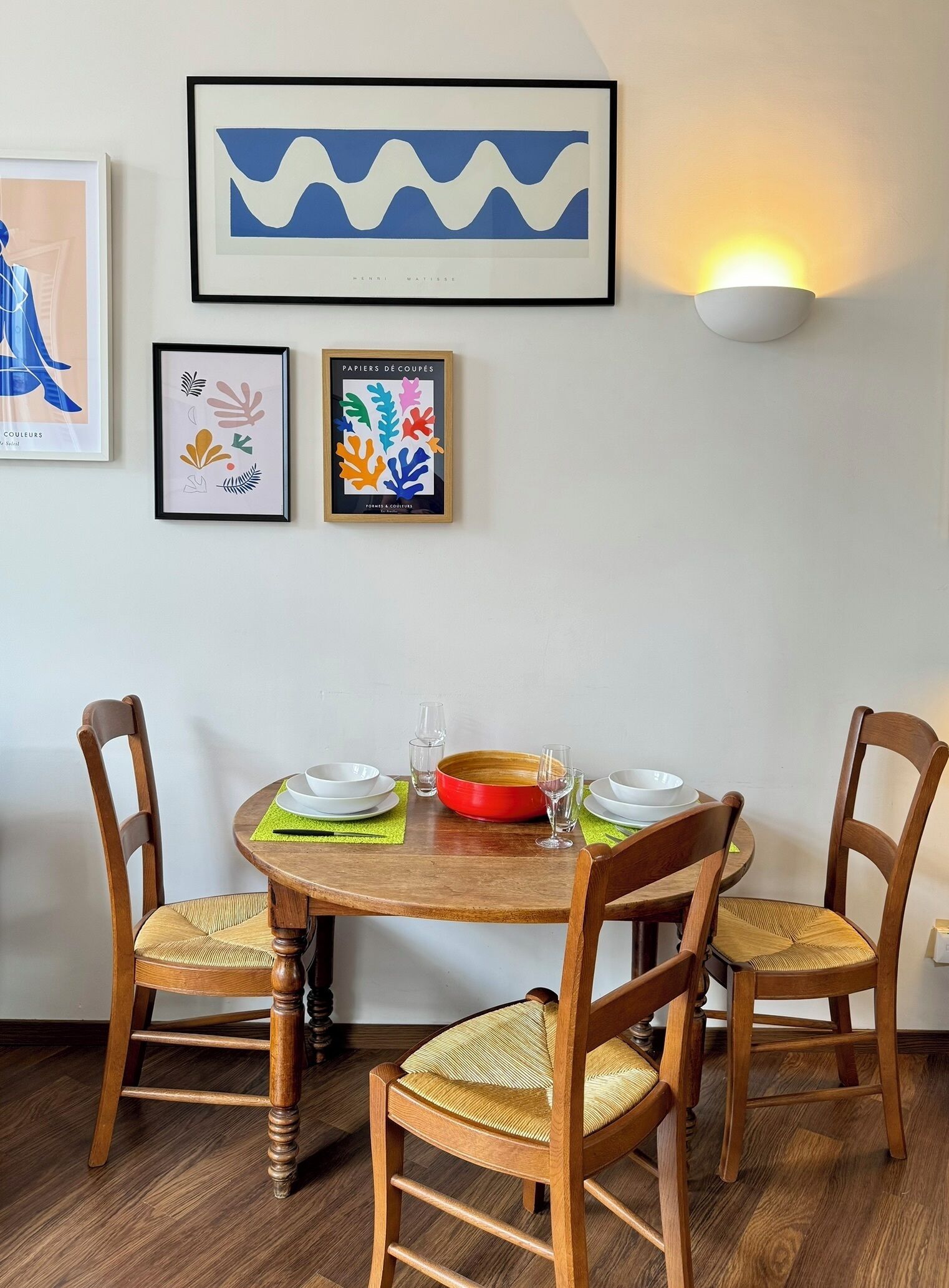 Un coin repas comprenant une table ronde en bois, quatre chaises en bois avec des assises en osier, et un luminaire fixé au mur.