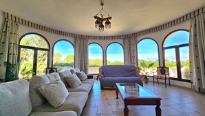 A spacious living room with a sofa, armchair, and coffee table, large arched windows with a view of greenery, and a chandelier. - Castell Villa Bohío Mallorca (Lluchmajor)