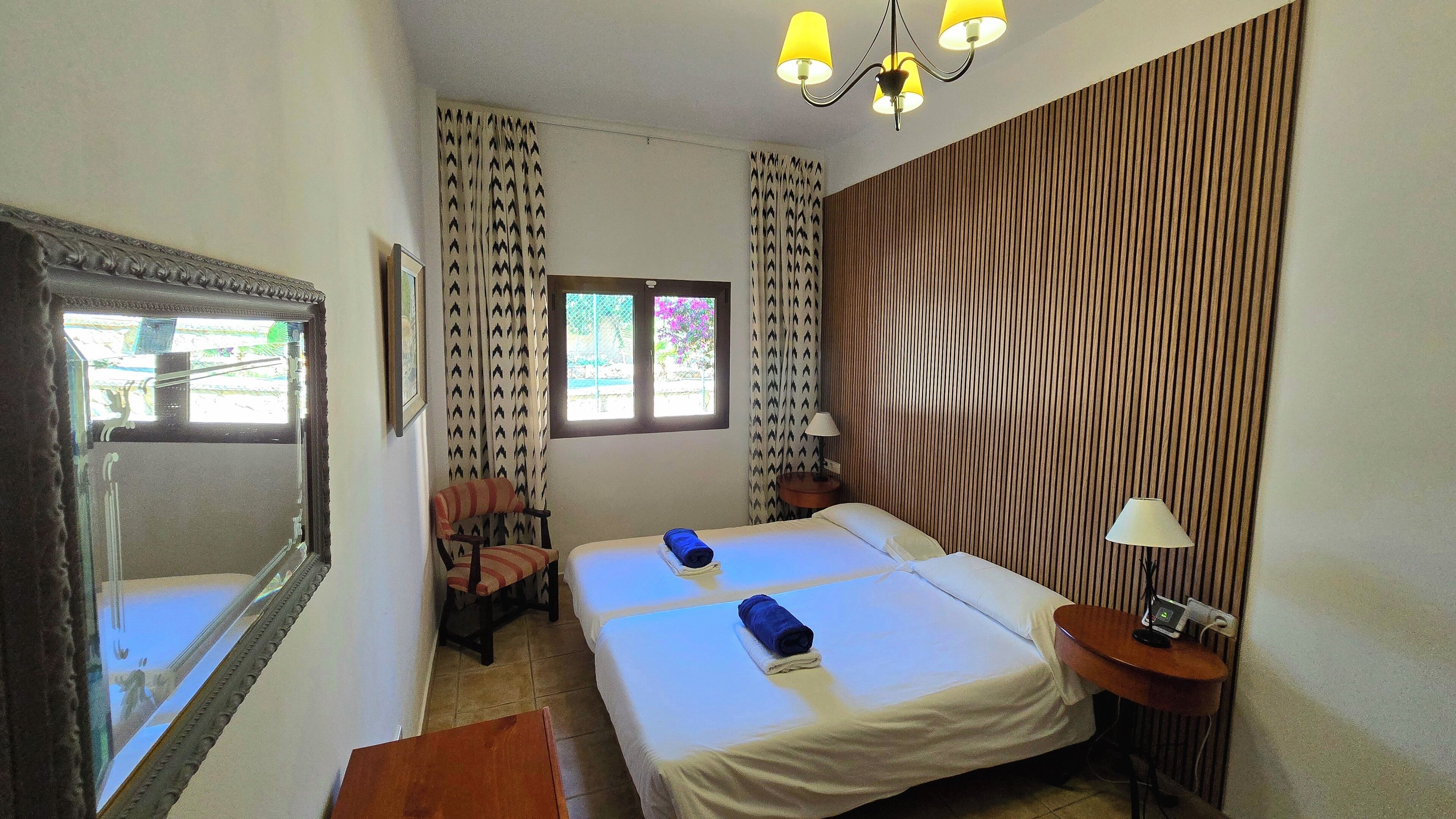 Una habitación de hotel con dos camas, un espejo grande, una ventana con cortinas, un panel de pared de madera y una mesita con una lámpara.