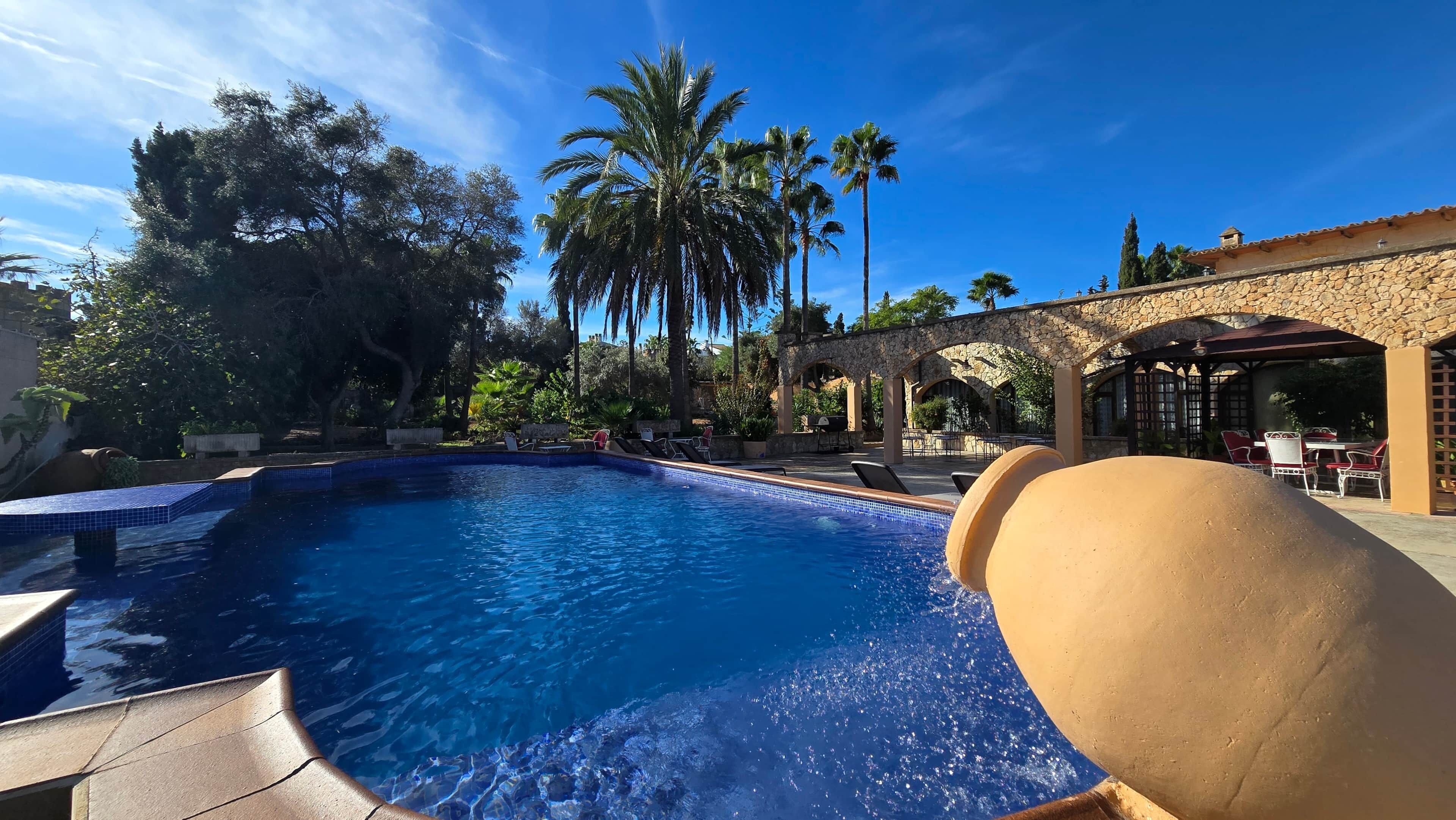 A swimming pool with clear blue water, surrounded by palm trees and a stone building with outdoor seating.