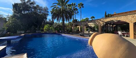 A swimming pool with clear blue water, surrounded by palm trees and a stone building with outdoor seating.