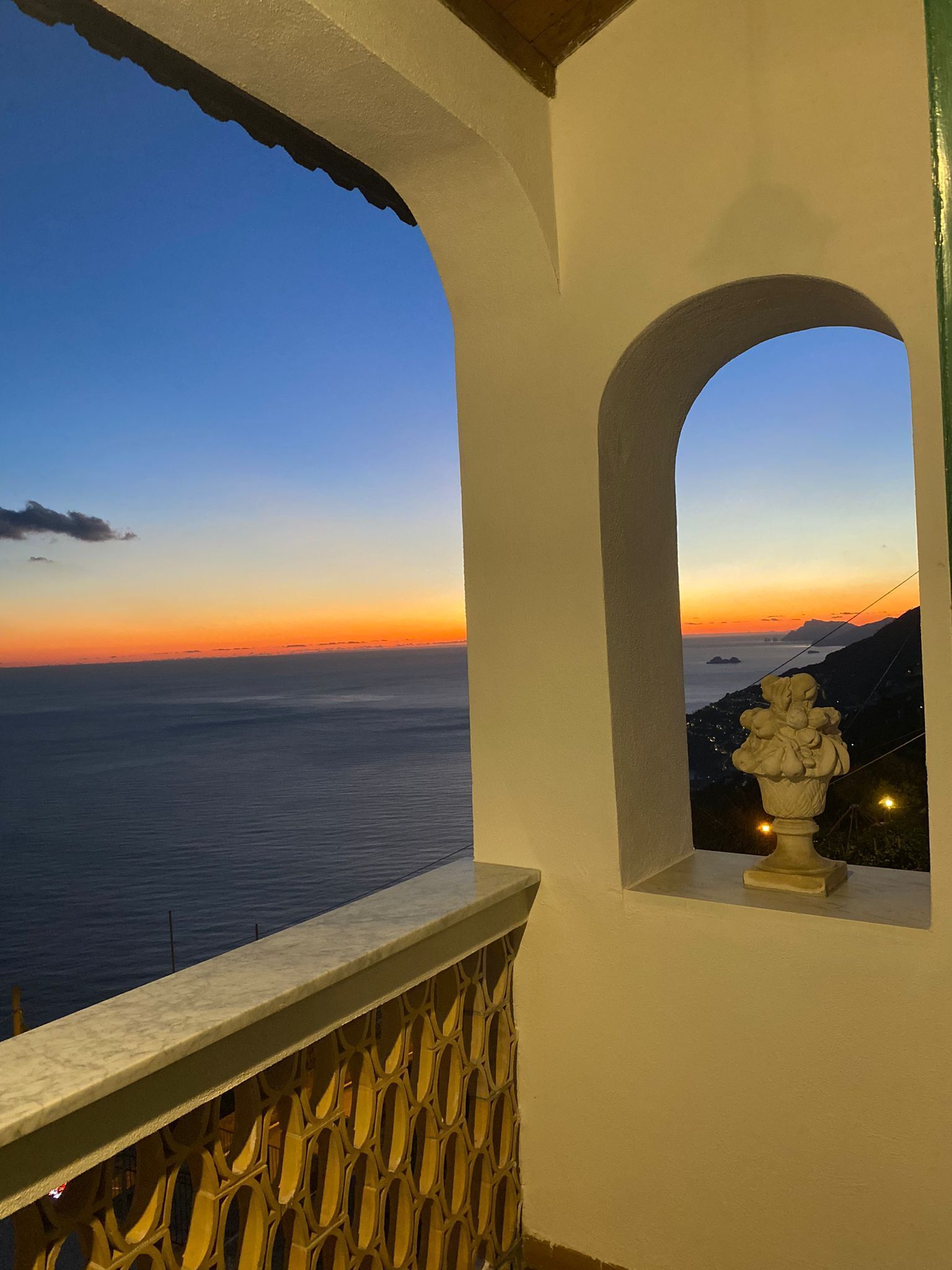 Ein Balkon mit Blick auf das Meer bei Sonnenuntergang, eine Ziervase und ein verzierter Geländer.