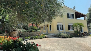 Um edifício amarelo de dois andares com um pátio pavimentado em pedra, cercado por vegetação exuberante e plantas floridas.