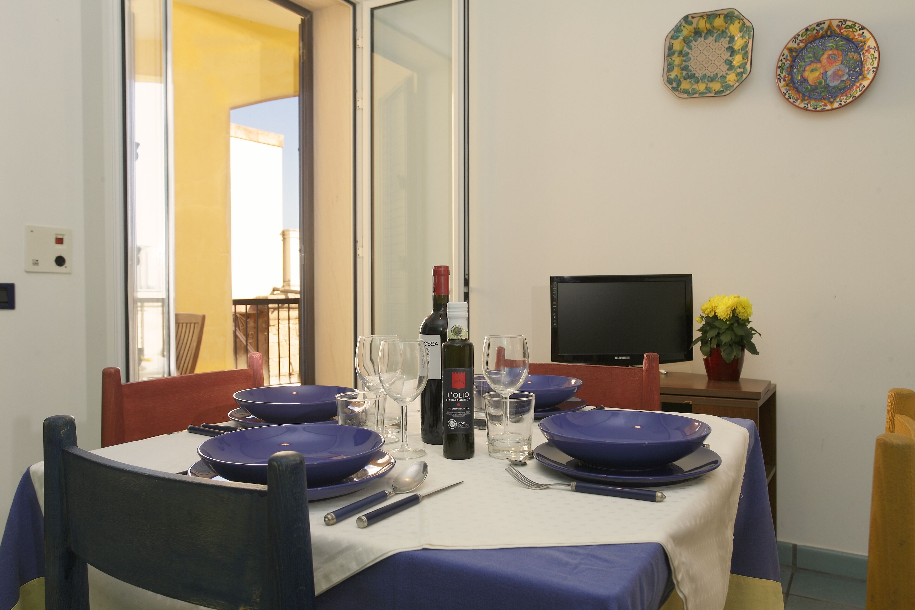 A dining table set with blue plates, wine glasses, and cutlery, with a bottle of wine and a television in the background.