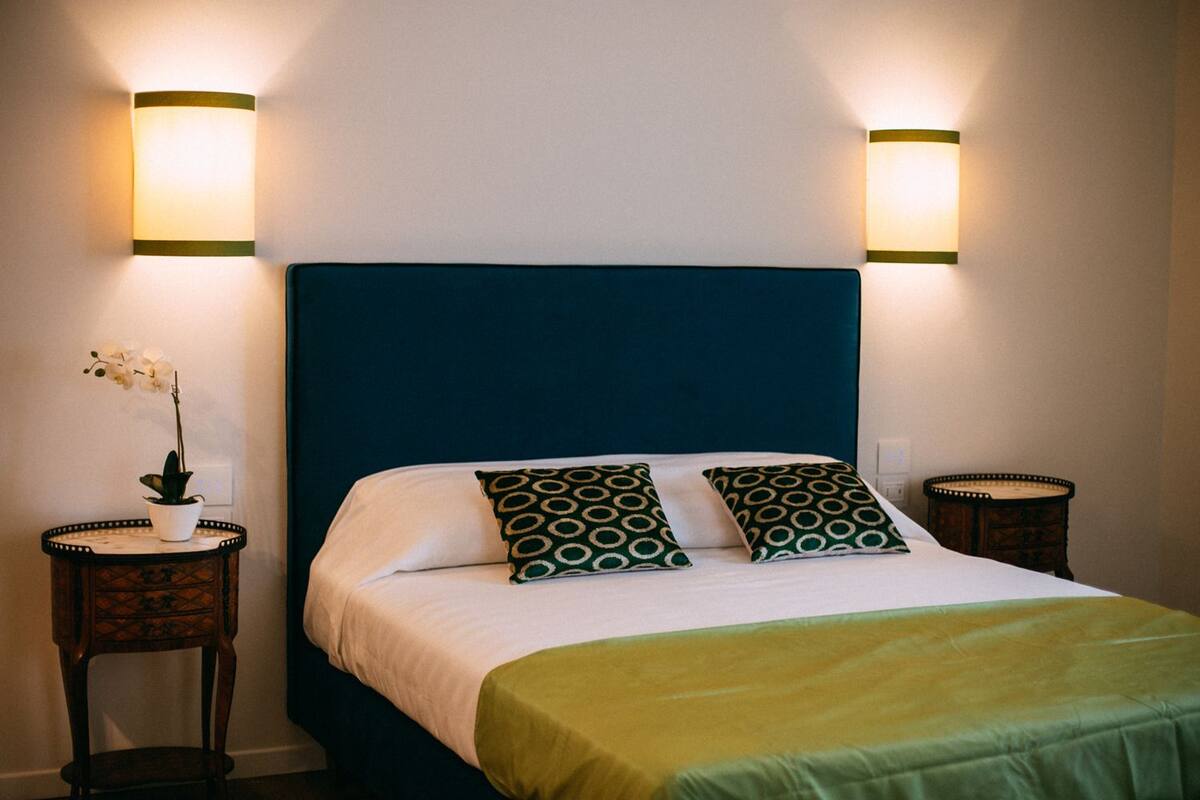 A neatly made bed with a green headboard, two patterned pillows, and a white comforter. There are two bedside tables with lamps, one of which has a potted orchid.
