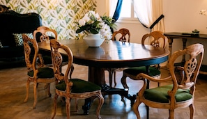 Un comedor con una mesa redonda de madera rodeada por seis sillas de madera con cojines verdes. Un jarrón blanco con flores blancas está colocado sobre la mesa.