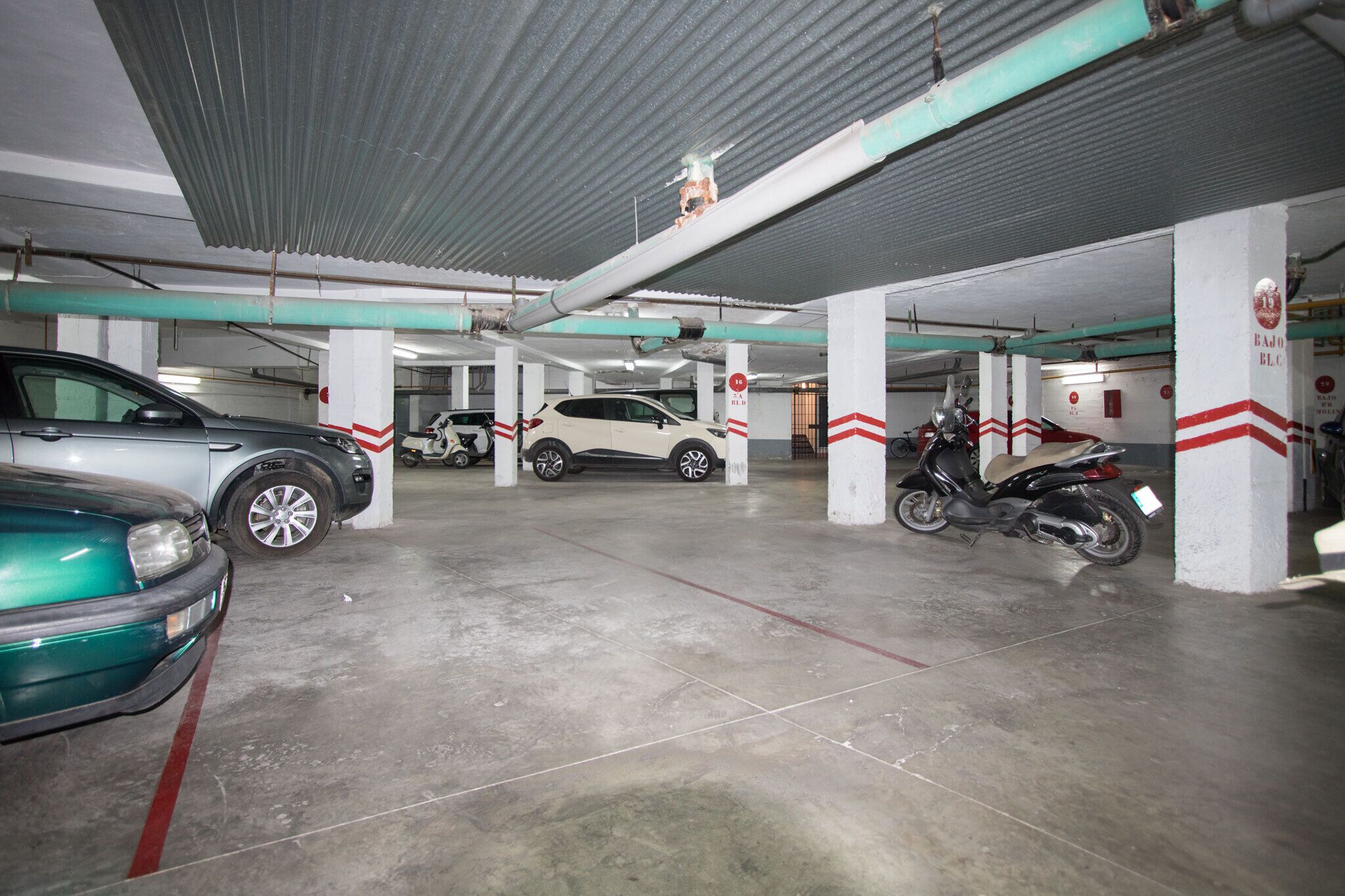 A multi-level parking garage with concrete floors and white pillars, featuring several parked cars and scooters.