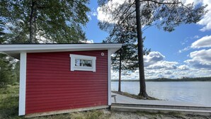 A red shed with a small window, a dock extending into a lake, and a dog lying on the ground. - Lillarmsjö-cottage, cottage with lake view. (Lillarmsjö)