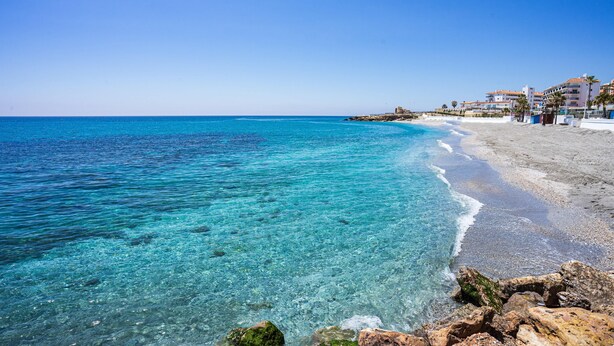 A beach with clear turquoise water, a rocky shoreline, and buildings with balconies in the background.