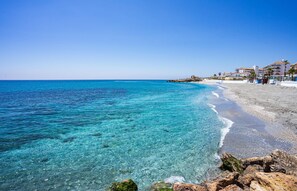Une plage à l’eau turquoise et limpide, un rivage rocheux et, au loin, des bâtiments dotés de balcons.