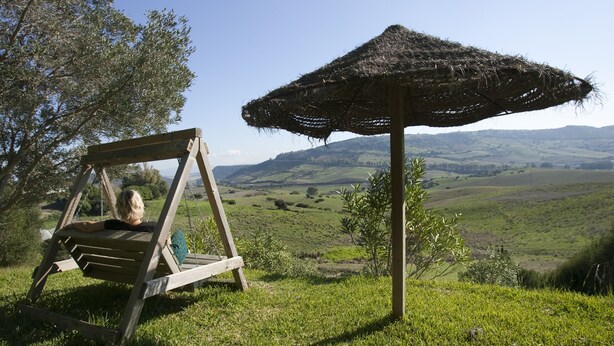 Une chaise à bascule en bois, placée sous un parasol en chaume, avec en arrière-plan des collines ondoyantes et un ciel dégagé.