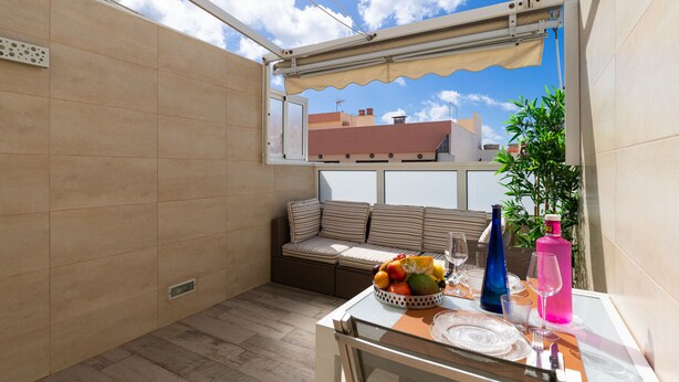 Eine sonnendurchflutete Terrasse mit bequemen Sitzgelegenheiten, einem Esstisch und Blick auf Himmel und Gebäude.