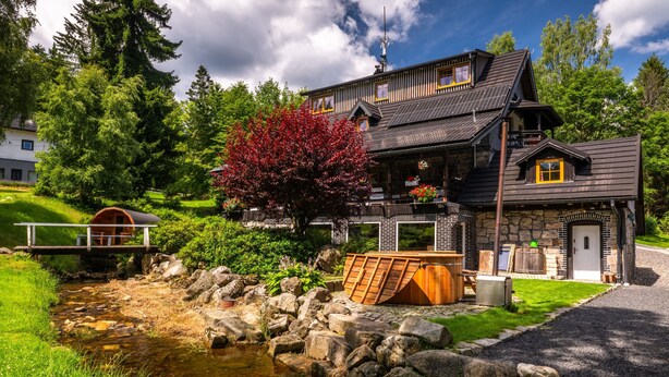 A wooden house with a stone foundation, a wooden deck, and a small pond.