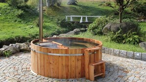 A wooden hot tub with a ladder, surrounded by a cobblestone patio and greenery.