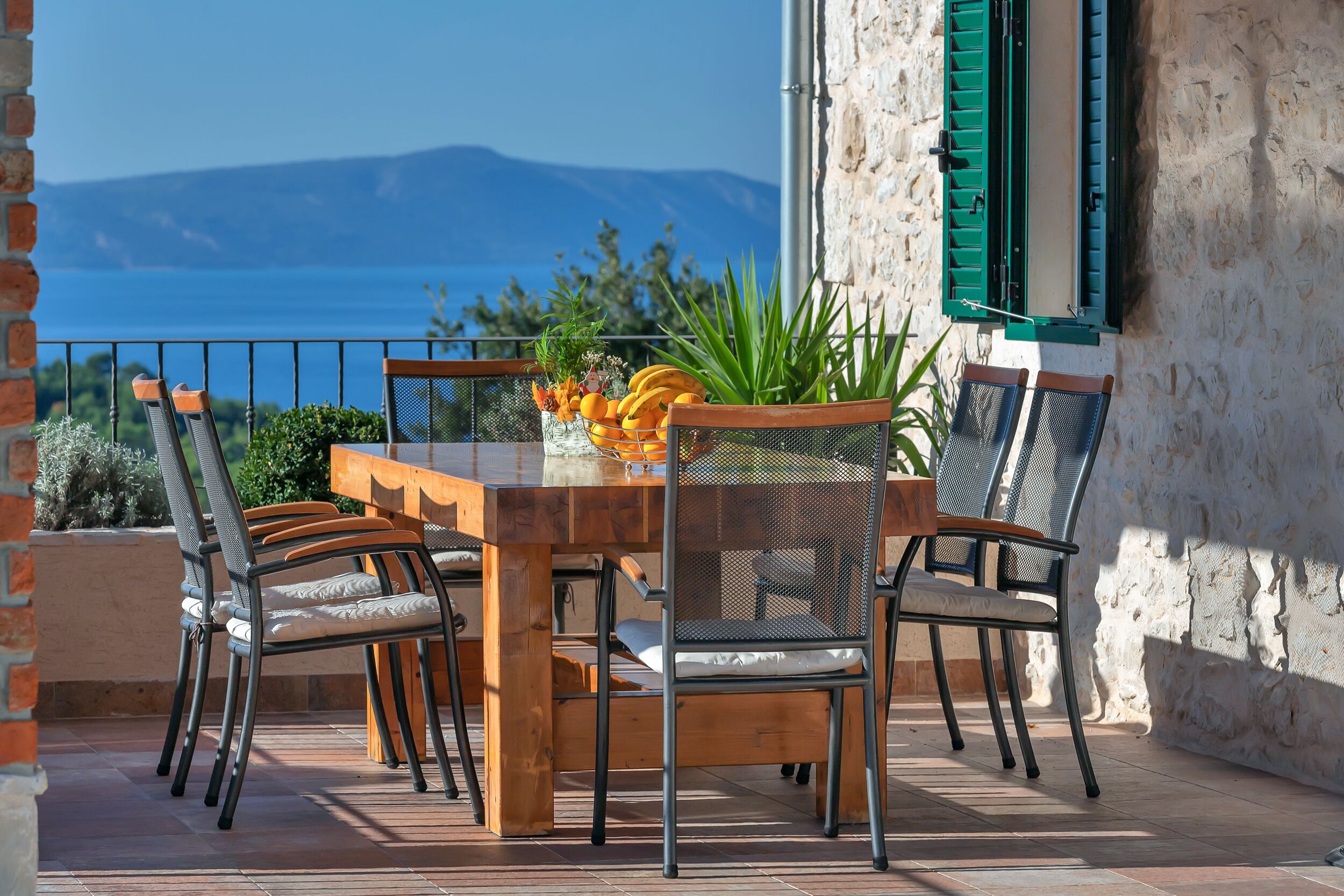 A wooden table with chairs, a vase of flowers, and a view of the sea and mountains.