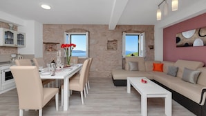 A modern living room with a beige sofa, a dining table with chairs, and a kitchen area with a stone wall and windows. - NEW! Stone house in quiet location, heated pool + sea views, ideal for familys (Zivogosce)