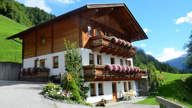Eine zweigeschossige Holzhütte mit Balkon, der mit Blumen geschmückt ist, einem kleinen Garten und freiem Blick auf die umliegenden Berge.