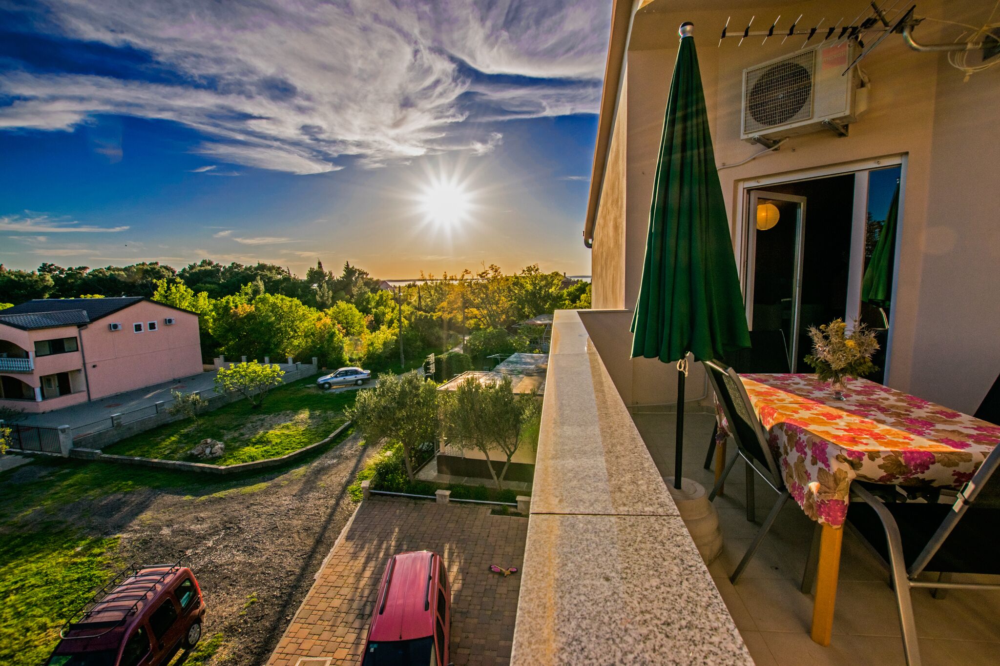 A balcony with a table and chairs, overlooking a residential area with a pink building and parked cars.