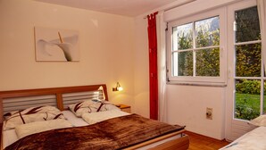 Une chambre à coucher avec un lit en bois, une table de chevet, une lampe fixée au mur et une fenêtre donnant sur un paysage verdoyant.