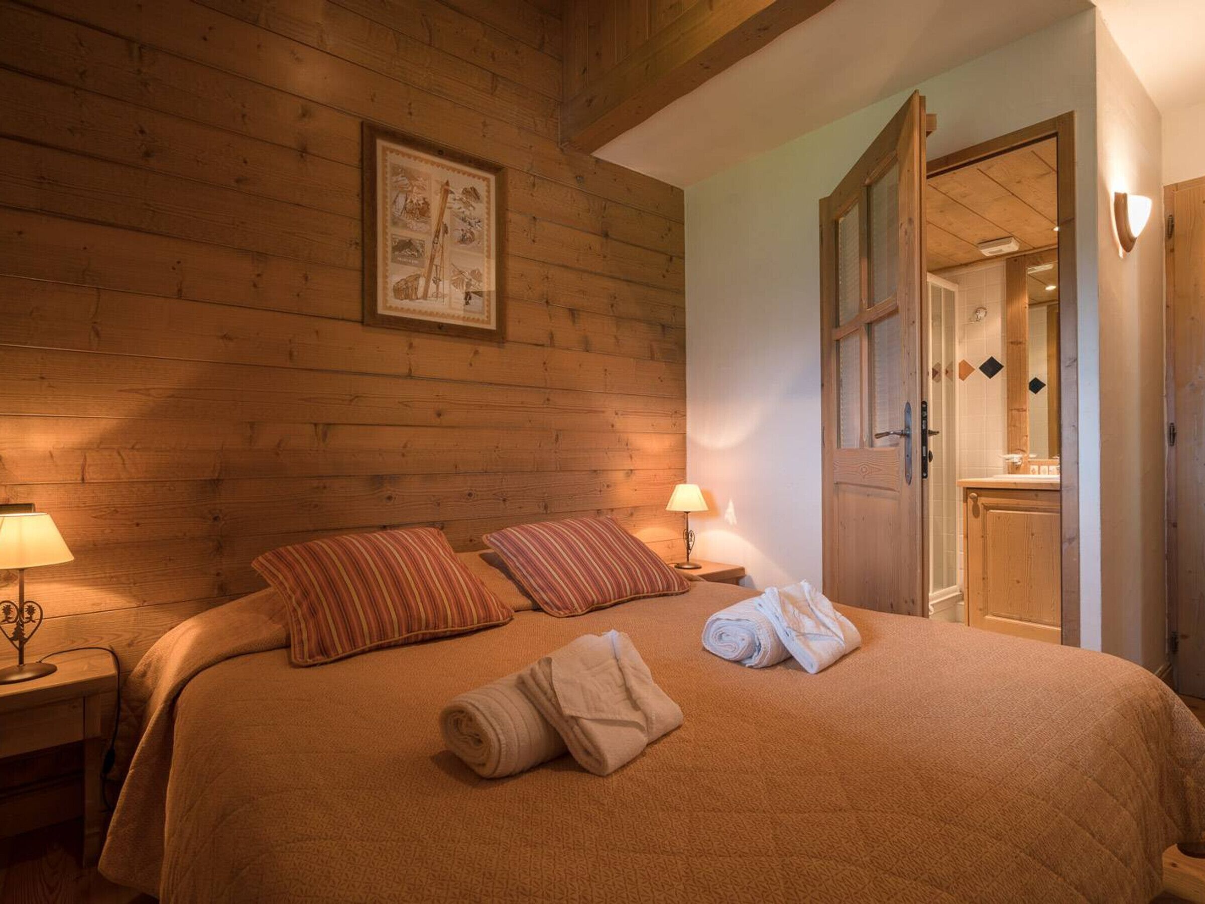 Ein gemütliches Zimmer mit Holzwänden, einem Bett mit gestreifter Bettwäsche, einem gerahmten Bild und einem durch eine offene Tür einsehbaren Badezimmer.