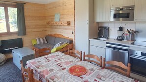 A cozy cabin-style room with a wooden interior, a dining table with a checkered tablecloth, a sofa, a small kitchenette with a microwave and stove, and a window with curtains. - Nice 3-bedroom, 2-bath apartment in Valfréjus, 6/8 people (Valfréjus)