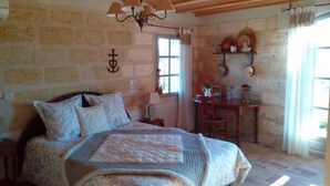 A bedroom with a stone wall, a bed with a patterned blanket, a chandelier, a window with curtains, and a small table with a chair. - Camargue Bed and Breakfast (Le Cailar)