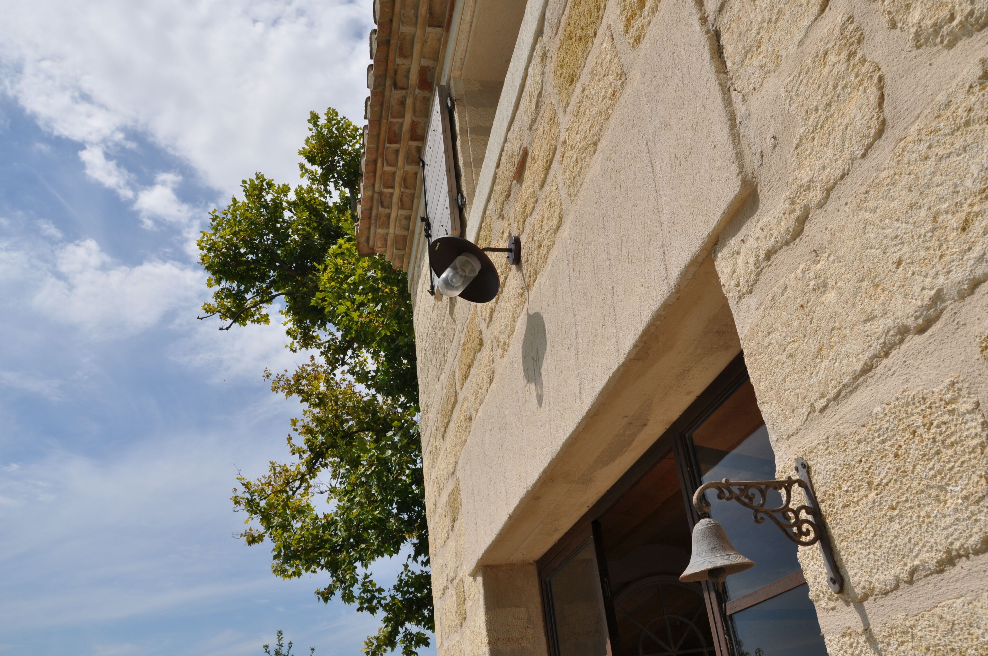 A building with a textured stone facade, a bell mounted on the wall, and a window with a view of the sky.