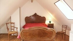 Un dormitorio con techo inclinado, una cama de madera, dos sillas, una mesita de noche con una lámpara y un radiador.