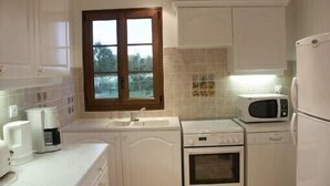 Cocina compacta con armarios blancos, microondas, cafetera y ventana con vista a árboles.