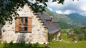 Una casa in pietra con persiane di legno, un giardino fiorito e montagne sullo sfondo.