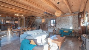 Un interno rustico con soffitto in legno, pareti in pietra, zona pranzo, soggiorno con divano e poltrone, e cucina con piano cottura.