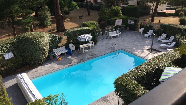 Une piscine entourée d’arbres, avec des chaises longues et des parasols, et un bâtiment visible au loin.