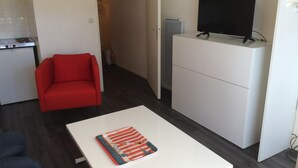 Un espace de vie compact comprenant un coin cuisine, un canapé rouge, une table blanche avec un livre et un téléviseur posé sur un meuble.
