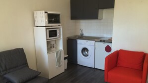 Une cuisine compacte équipée d’un four à micro-ondes, d’une machine à café et d’un lave-linge. Le salon comprend un canapé gris et un fauteuil rouge.