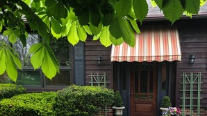Ein Haus mit Holzfassade, einem gestreiften Sonnenschirm, einer Haustür und einem kleinen Garten mit einem Blumentopf und einem Busch.