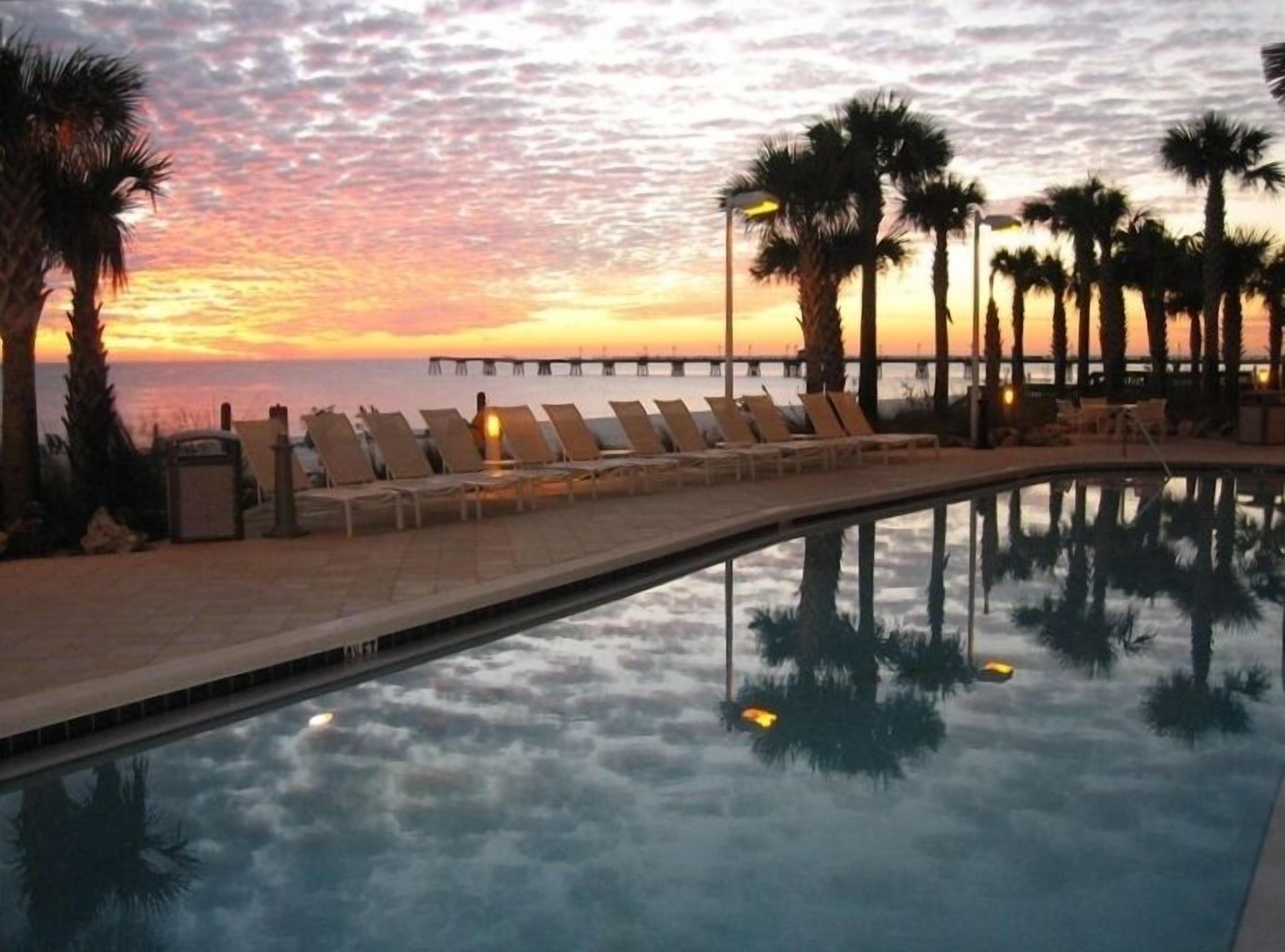 A poolside area with lounge chairs and palm trees, overlooking a body of water at sunset.