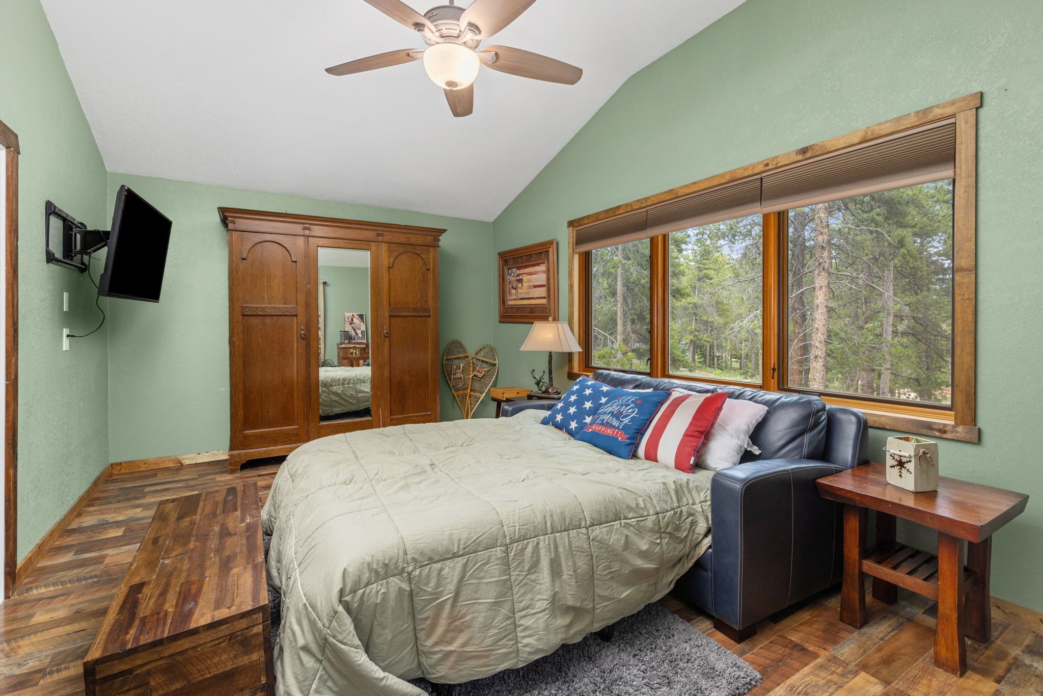 A bedroom with a bed, a ceiling fan, a window with a view of trees, a wooden wardrobe, and a television mounted on the wall.