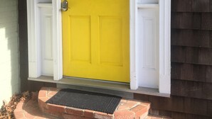 Une porte jaune, un porche blanc et un petit escalier en briques.
