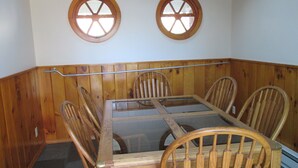 Un coin repas avec une table à plateau de verre entourée de chaises en bois, deux fenêtres à cadre en bois et un mur avec un lambris en bois.